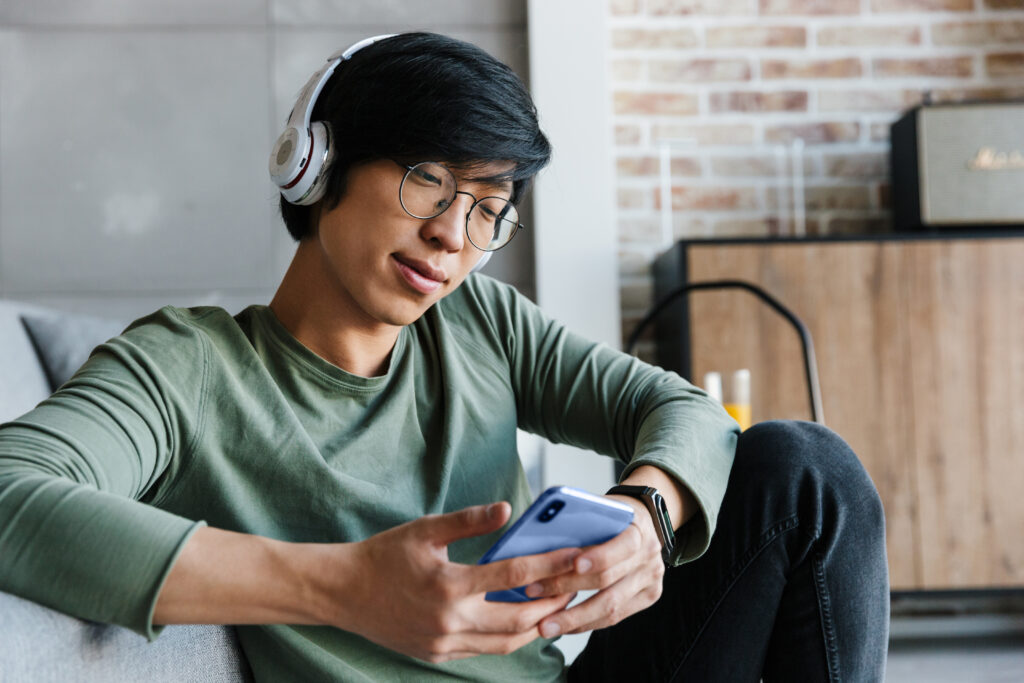 Image of concentrated asian man in eyeglasses using cellphone and wireless headphones while sitting in living room